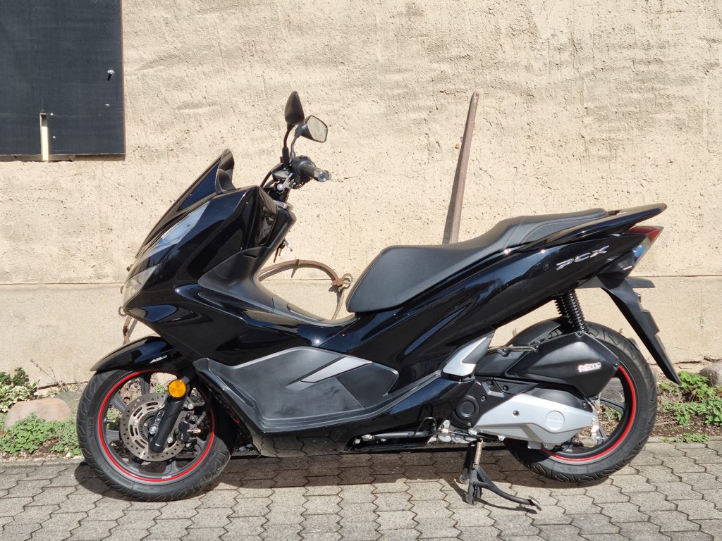 Schwarzer Honda PCX Motorroller in der Seitenansicht, gepflegter Zustand mit roten Felgenrandaufklebern auf Kopfsteinpflaster vor einer hellen Wand
